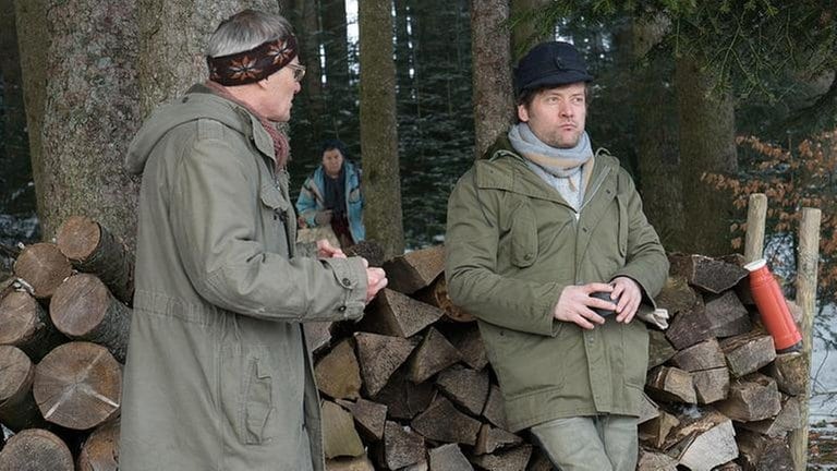 Karl und Riedle machen Pause von der Waldarbeit, im Hintergrund sieht man Lioba, die die beiden belauscht