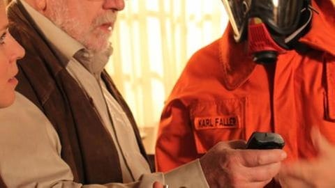 Bea und Hermann stehen neben Karl, der in Feuerwehrausrüstung seinen Alarmpiepser checkt