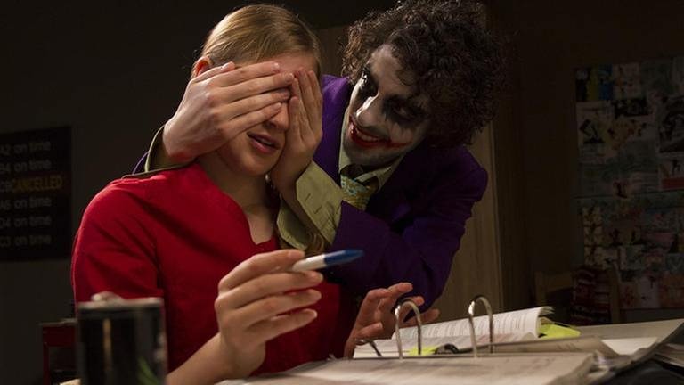 Albert, als Joker verkleidet, hält Celine die Augen zu