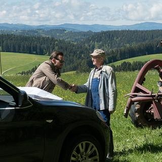 Constantin und Karl begrüßen sich auf freiem Feld, im Vordergrund Constantins Limousine, daneben Karls Traktor