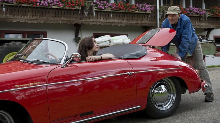 Sabine sitzt in ihren Porsche-Oldtimer, Karl hat die Motorhaube geöffnet und versucht etwas zu reparieren