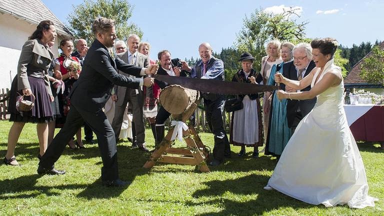 Eva und Andreas zersägen einem alten Hochzeitsbrauch nach einen Baumstamm, die Hochzeitsgesellschaft schaut und jubelt zu