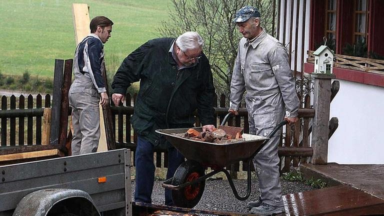 Karl schiebt eine Schubkarre voller alter Backsteine, aus der Hermann sich einen nimmt, im Hintergrund hantiert Sebastian mit einem langen Brett