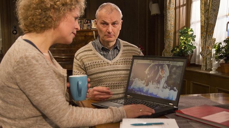 Bea und Karl sitzen am Wohnzimmertisch, Bea arbeitet am Laptop