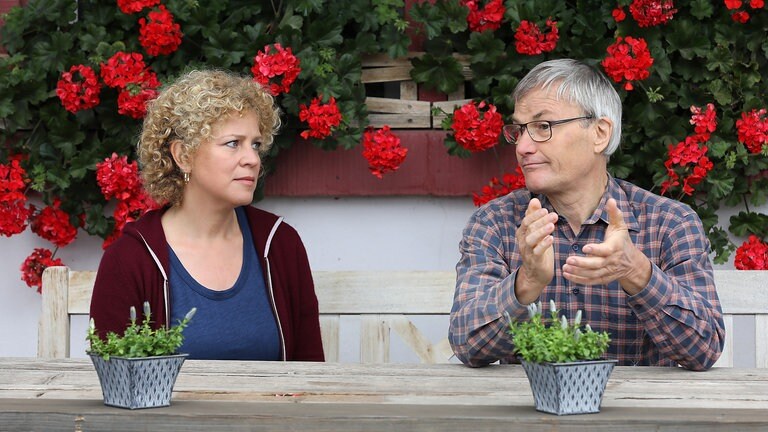 Bea und Karl sitzen auf einer Bank vor dem Hof, im Hintergrund Geranien