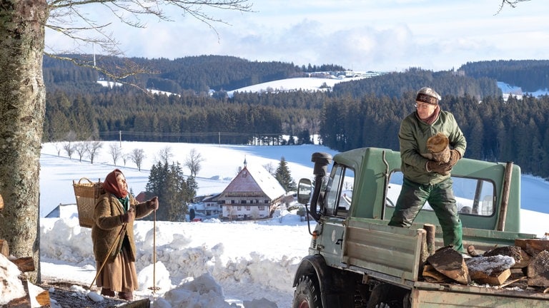 Karl steht auf seinem Unimog und lädt Holz ab, Lioba kommt durch den Schnee gelaufen