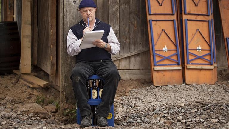 Toni sitzt mit Block und Bleistift auf einer Bierkiste vor der renovierten Mühle