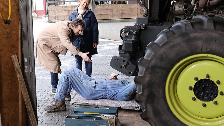 Karl liegt unter seinem Traktor, davor stehen Bea und ein Feriengast, der Karl begrüßen möchte