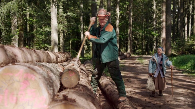 Karl entrindet einen Baum, Lioba kommt den Waldweg entlang auf ihn zu gelaufen.