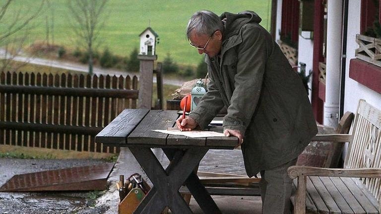 Karl steht am Tisch vor dem Fallerhof und schreibt etwas auf