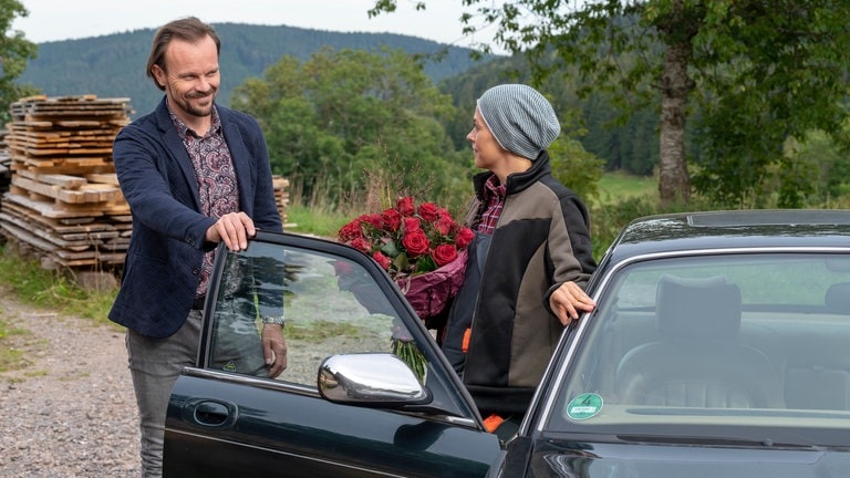 Constantin hält Sophie, die einen Strauß roter Rosen in der Hand hat, die Autotür auf