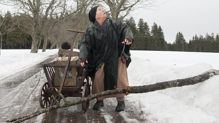 Lioba steht in schneebedeckter Landschaft und guckt in den Himmel, vor ihr auf dem Boden liegt ein dicker Ast