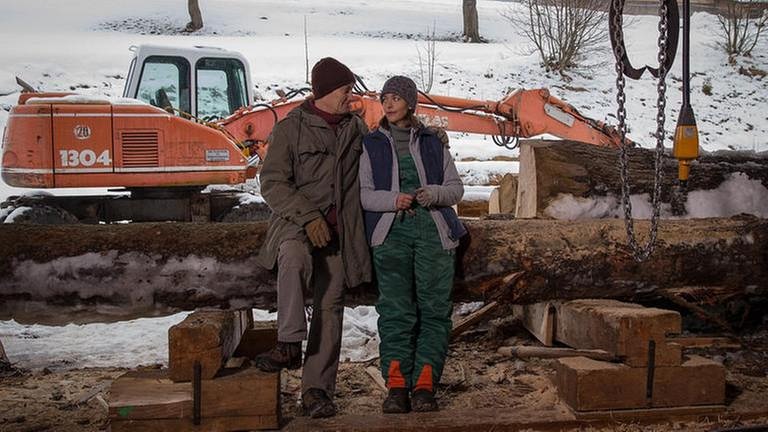 Karl und Sophie sitzen im Sägewerk und unterhalten sich, im Hintergrund ein orangefarbener Bagger