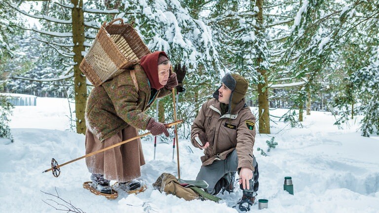 Sebastian kniet im Schnee, Lioba steht neben ihm, sie trägt einen Weidekorb auf dem Rücken