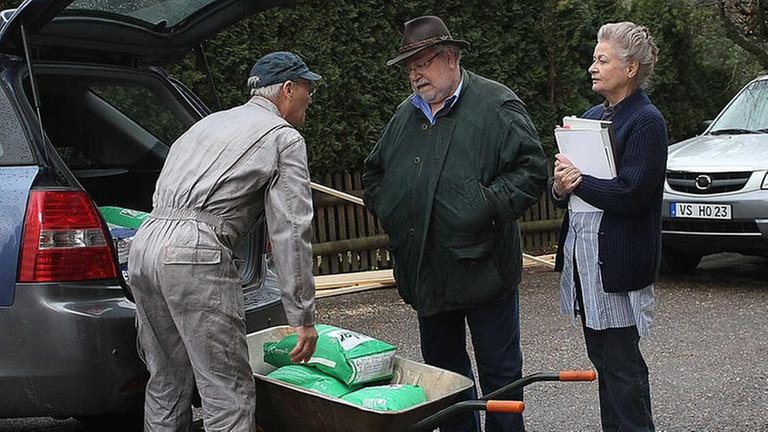 Karl entlädt Betonsäcke aus seinem Auto, Hermann und Johanna stehen dabei und unterhalten sich mit ihm