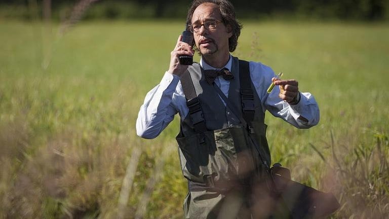 Herr Helmhorst (Christoph Sonntag), der Wasserspezialist steht auf einer Wiese und telefoniert, in der linken Hand ein Reagenzglas mit einer Wasserprobe