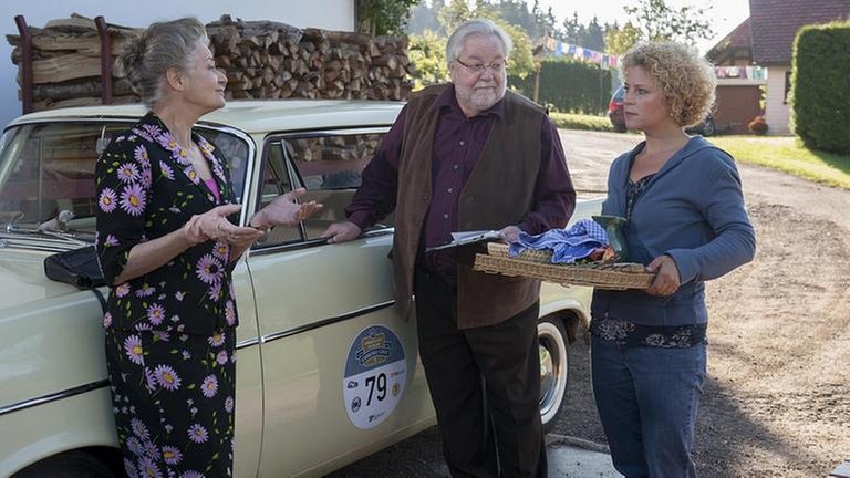 Johanna und Hermann stehen vor ihrem Oldtimer, Bea kommt mit einem Tablett in den Händen dazu