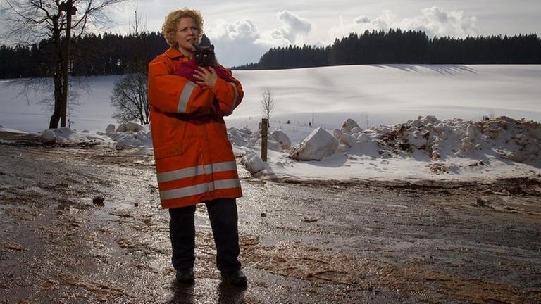 Bea steht in Feuerwehrmontur in Schneelandschaft und hat eine in eine Decke gewickelte schwarzgraue Katze auf dem Arm