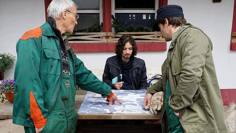 Karl, Albert und Riedle besprechen einen Plan, der auf dem Holztisch vor dem Fallerhof liegt