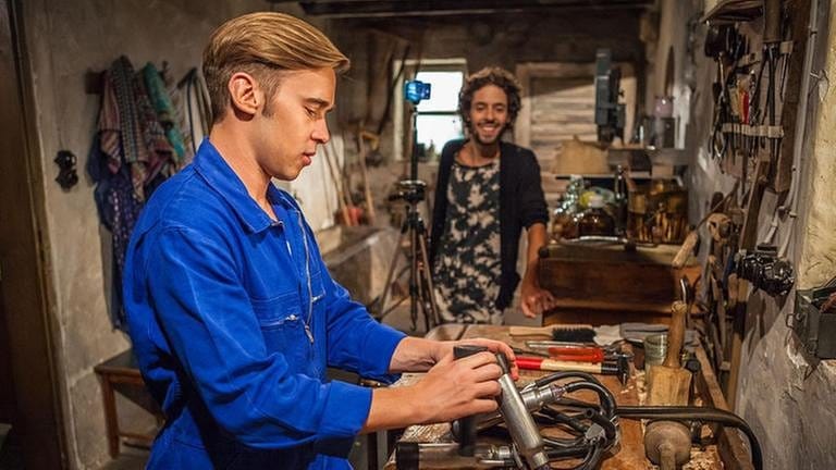 Sebastian arbeitet an der Werkbank, im Hintergrund sieht man Albert mit seiner Handykamera