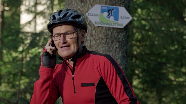 Karl steht in Fahrradmontur mit Helm im Wald und telefoniert