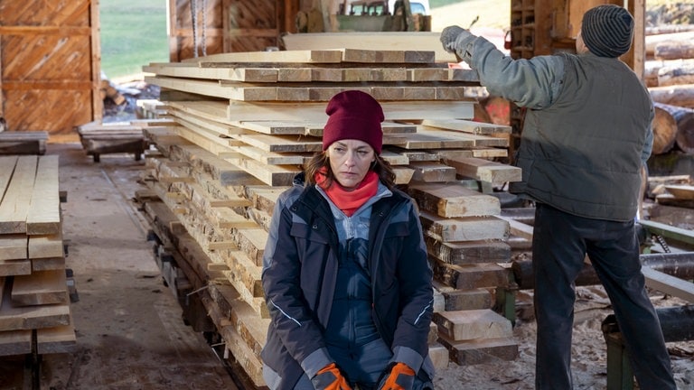 Sophie (Janina Flieger) sitzt auf einem Stapel Holz im Sägewerk, Niki (Niki König) arbeitet