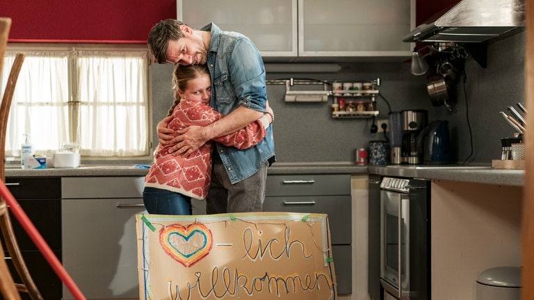 Carlotta (Mila Breiter) und Andreas (Ralph Gassmann) umarmen sich. Vor ihnen steht ein Willkommens-Schild für Eva.