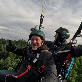 Zwei Männer sind an einem Fallschirm in der Luft und zeigen Daumen nach oben