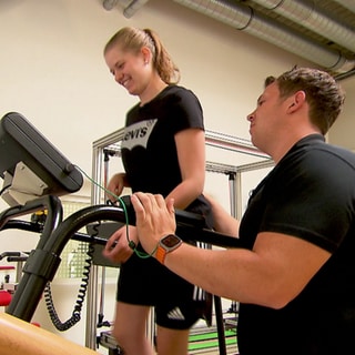 Eine junge Frau mit Parkinson steht auf einem Laufband mit einem Trainer an der Seite. 