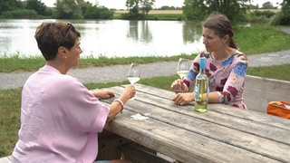 Die Reporterin und die Ex-Weinkönigin sitzen auf einer Bank mit Tisch und trinken Wein