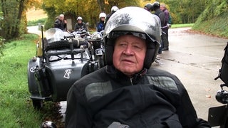 Adolf Mühlich sammelt historische Motorräder. Er trägt Motorradkluft und steht vor einem Motorrad.