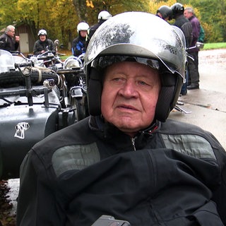 Adolf Mühlich sammelt historische Motorräder. Er trägt Motorradkluft und steht vor einem Motorrad.