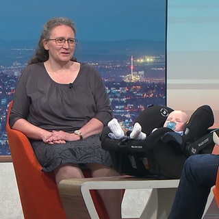 Mann und Frau Werner sitzen im Studio. Ihr Baby schläft in einer Babytrage. 