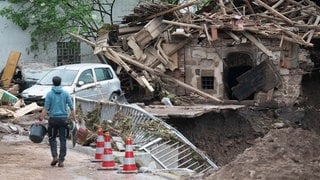 Ein von Hochwasser stark zerstörtes Gebäude neben Erdrutsch, davor ein Mann mit Eimer.