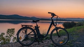 Ein E-Bike steht vor einem See bei Sonnenuntergang. Akkus: Brandgefahr und Entsorgung.