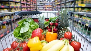 Ein Einkaufskorb mit Obst und Gemüse steht in einem Supermarkt. 