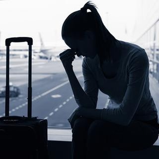 Eine Frau sitzt neben einem Koffer, verzweifelt mit dem Kopf auf eine Hand gestützt, vor einem Fenster am Flughafen.