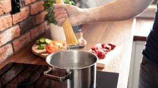 Spaghetti werden in einen Topf heißes Wasser gegeben. Im Hintergrund sind weitere Zutaten für ein Nudelgericht zu sehen. Tipps zum Nudelkochen erfahren Sie bei uns.