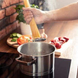 Spaghetti werden in einen Topf heißes Wasser gegeben. Im Hintergrund sind weitere Zutaten für ein Nudelgericht zu sehen. Tipps zum Nudelkochen erfahren Sie bei uns.