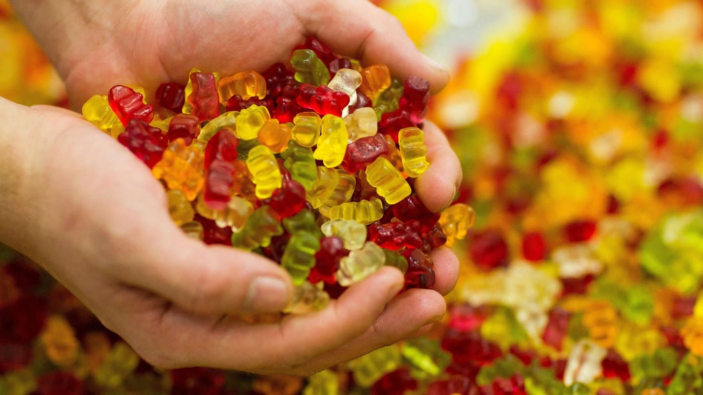 Zwei Hände halten eine handvoll bunter Gummibärchen. Im Hintergrund ist weitere zuckerhaltige Fruchtgummi-Süßigkeiten zu sehen.