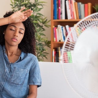 Frau sitzt schwitzend mit einer Hand an der Stirn vor einem weißen Ventilator in ihrer Wohnung, um sie abzukühlen. Die Sommer werden heißer, Wohnungen und Häuser unerträglich warm. Mit diesen Tricks bekommen Sie Ihre Wohnung ohne Klimaanlage kühl!