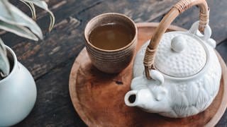 Braune Tasse mit Tee und eine weiße Kanne mit Henkel auf einer braunen Platte. Links sieht man eine Pflanze in einem weißen Topf. Die Gegenstände liegen auf einem dunkelbraunen Tisch aus Holz