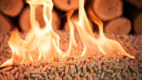 Pellets aus Sägespänen stehen in Flammen. Im Hintergrund sind Holzbalken aufeinander gestapelt.