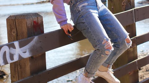 Mädchen mit zerschlissenen Jeans sitzt auf einem Zaun. Jeans im Used Look schaden in der Herstellung der Umwelt. Geht das auch nachhaltig?