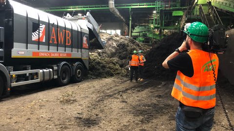 Am linken Bildrand entlädt ein Müllwagen Biomüll auf eine Halde in einer großen Halle. Im Vordergund steht ein Kameramann, der Ökochecker Tobias Koch und Kompostwerk-Sprecher Tilo Dumuscheit neben dem Müllwagen filmt. Am rechten Bildrand erkennt man eine Treppe die zu den Verarbeitungsanlagen führt.