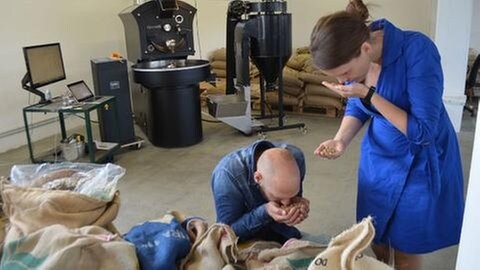 Ein Mann und eine Frau halten grüne Kaffeebohnen aus großen Säcken  in den Händen und schnuppern daran