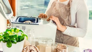 Eine Person steht an einem Brockbackautomaten und hat gerade den Teig für ein Brot reingelegt. Im Vordergrund stehen einige Backzutaten.