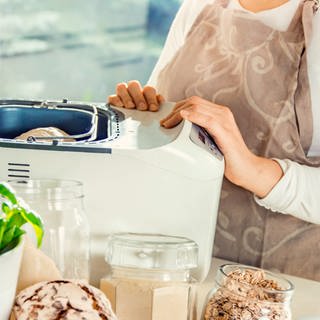 Eine Person steht an einem Brockbackautomaten und hat gerade den Teig für ein Brot reingelegt. Im Vordergrund stehen einige Backzutaten.