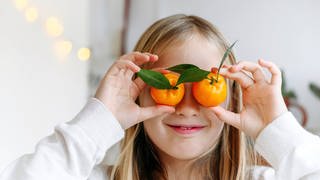 Ein blondes Mädchen hält sich lächelnd zwei Mandarinen vor die Augen: Eine gesunde, vitaminreiche Ernährung ist wichtig für die Augen