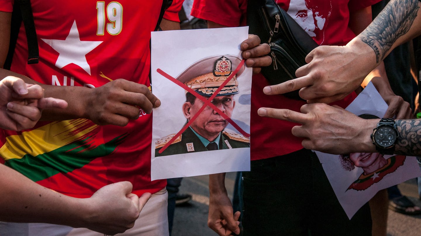 Demonstranten halten ein Porträt von Armeechef Min Aung Hlaing, über dessen Gesicht ein rotes Kreuz gemalt wurde. Eine zeigen mit dem Mittelfinger auf das Bild: Das Militär hatte am 1. Februar 2021 die zivile Führung Myanmars um Aung San Suu Kyi durch einen Putsch entmachtet und einen einjährigen Ausnahmezustand verhängt. Das Wort 